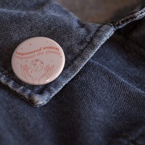 May include: A pink button with the text "empowered women empower the planet" in red. The button features a line drawing of a globe with hands reaching up to hold it.