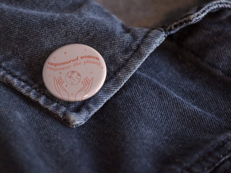May include: A pink button with the text "empowered women empower the planet" in red. The button features a line drawing of a globe with hands reaching up to hold it.