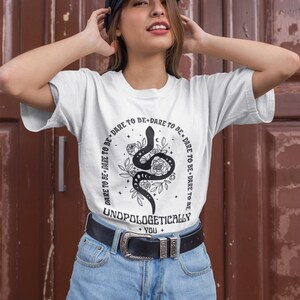 Pu&ograve; includere: Maglietta bianca con un serpente nero e un disegno floreale. Il testo sulla maglietta recita "DARE TO BE+DARE TO BE DARE TO BE+DARE TO BE UNDPOLOGETICALLY + YOU +"