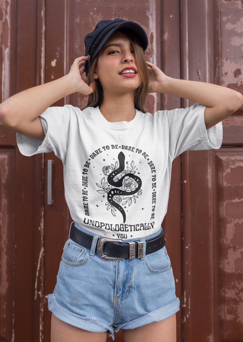 Pu&ograve; includere: Maglietta bianca con un serpente nero e un disegno floreale. Il testo sulla maglietta recita "DARE TO BE+DARE TO BE DARE TO BE+DARE TO BE UNDPOLOGETICALLY + YOU +"