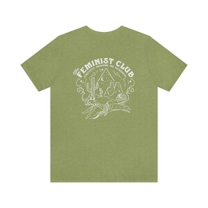 May include: A green t-shirt with a white graphic design on the back. The design features a cactus, mountains, a crescent moon, and stars. The text reads "The Feminist Club: Tirelessly Smashing the Patriarchy. We'll Literally Forever."