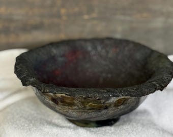 以下が含まれることがあります： 黒色のセラミックボウルで、縁が不均一でざらざらしています。ボウルは、黒に近い暗い釉薬で、赤と緑の色合いが見て取れます。