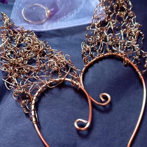 May include: Copper-coloured wire ear cuffs with intricate, abstract designs. The ear cuffs feature a delicate, woven pattern that extends upwards. A matching ring and other jewellery are also visible in the image. The jewellery is set against a dark background.