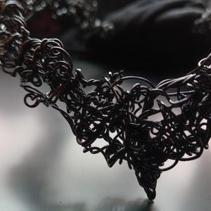 May include: A close-up of a dark, wire-like necklace with a complex, tangled design. The necklace is black and has a pointed bottom. The intricate design suggests a handcrafted piece of jewellery. The overall aesthetic is artistic.