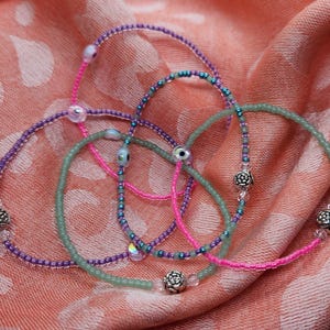 May include: A collection of beaded bracelets in shades of purple, pink, and green, with silver-colored floral accents. The bracelets are arranged on a peach-colored fabric with a white pattern, creating a soft and feminine aesthetic.
