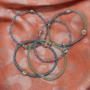 May include: Several beaded bracelets in shades of teal, green, and pink are arranged on a peach-colored fabric. The bracelets feature small beads and decorative silver accents. Some bracelets have small, round, gold-colored beads.