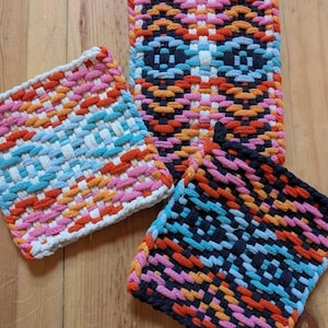 May include: Three colorful woven mats in shades of pink, orange, blue, and black. The mats are rectangular and square, with a textured, patterned design. The mats are made of a thick, durable material, and are likely used as trivets or placemats.