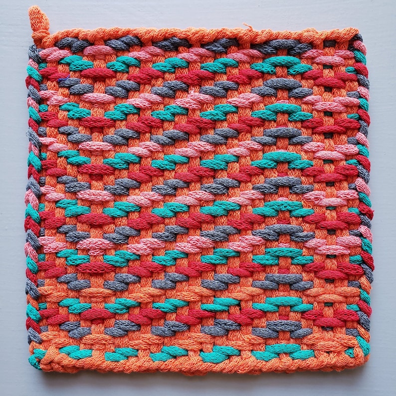 Puede incluir: Un agarrador de olla tejido con un patr&oacute;n geom&eacute;trico colorido en tonos naranja, rosa, turquesa y gris.