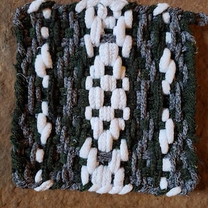 May include: A square, woven potholder with a central white design on a dark green and grey background. The potholder is made of thick yarn and has a loop for hanging. The texture is visible, showcasing the handmade quality.