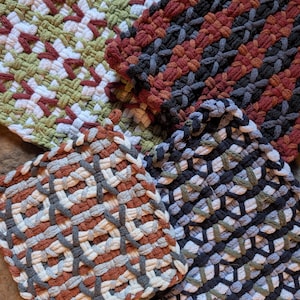 May include: Four square potholders made of woven cotton loops. One features olive green, white, and maroon. Another has dark charcoal and rust red. A third displays rust, charcoal, and white. The fourth shows charcoal, light blue, and white.
