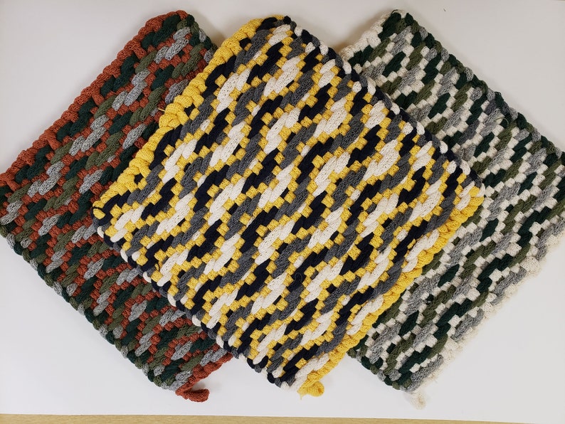 Puede incluir: Tres agarraderas de olla tejidas en diferentes patrones de colores. La primera es un patr&oacute;n marr&oacute;n, verde y gris. La segunda es un patr&oacute;n amarillo, negro, blanco y gris. La tercera es un patr&oacute;n verde, gris y blanco.