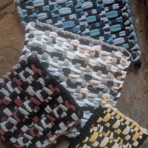 May include: A collection of woven potholders in various colors. The potholders feature a woven design with black, blue, gray, white, red, and yellow threads. Each potholder has a black border.