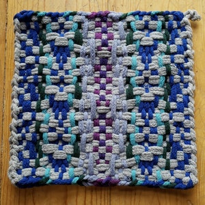 Może przedstawiać: Tkany podkładka pod gorące z szarym tłem i niebieskimi, zielonymi i fioletowymi paskami. Podkładka jest wykonana z bawełnianej przędzy i ma frędzlowany brzeg.