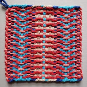 Puede incluir: Un agarrador de olla tejido con un patr&oacute;n de color rojo, rosa, azul y crema. El agarrador de olla tiene un lazo para colgar.