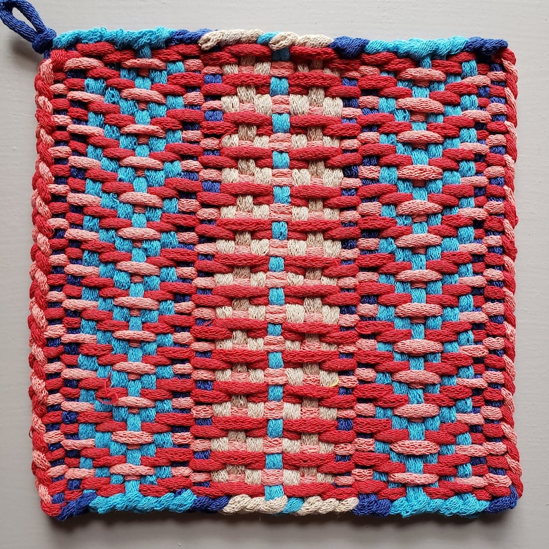 Puede incluir: Un agarrador de olla tejido con un patr&oacute;n de color rojo, rosa, azul y crema. El agarrador de olla tiene un lazo para colgar.