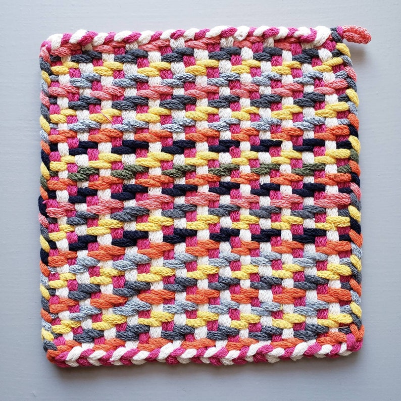 Puede incluir: Un agarrador de olla tejido de colores con un patr&oacute;n rosa, amarillo, naranja, gris y negro. El agarrador de olla est&aacute; hecho de hilo grueso tejido y tiene un borde blanco.