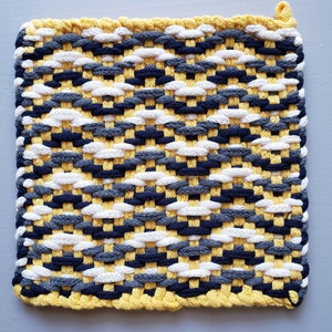 Puede incluir: Un posavasos tejido amarillo, blanco, gris y negro con un borde de flecos.