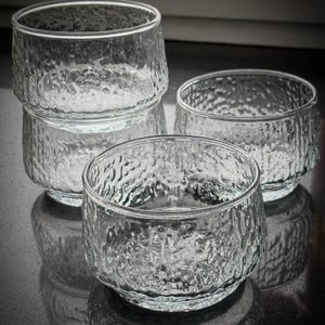 May include: Four clear glass bowls with a textured, hammered effect. The bowls are stacked and arranged on a reflective surface, highlighting their design. The glass has a slightly irregular rim.