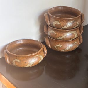 May include: Four round brown ceramic bowls with two small side handles each. The bowls feature a speckled texture and a decorative swirling pattern in a lighter tan colour around the exterior. One bowl sits alone while three are stacked vertically.