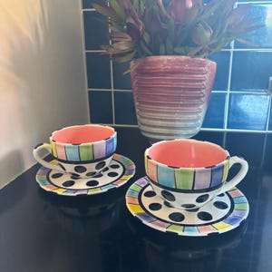 May include: Two ceramic teacups and saucers. The cups have striped rims in various colours and black polka dots. The interior of the cups is coral. The saucers and cups have a white base. A decorative vase with a plant is in the background.