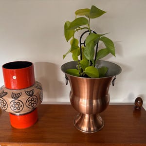 May include: A cylindrical ceramic vase with a bright orange top and bottom section and a beige middle band featuring a dark brown floral pattern, next to a metallic copper-coloured pedestal planter containing a green leafy plant.