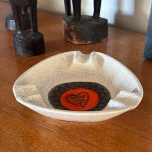 May include: A speckled, light grey ashtray with a central oval design. The oval features a red heart-shaped design within a dark brown border. The ashtray is on a wooden surface, with carved wooden figures in the background.