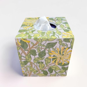 May include: A square tissue box cover featuring a botanical pattern of yellow honeysuckle flowers and green leaves on a light grey background. A single white tissue is visible protruding from the circular opening at the top of the box.