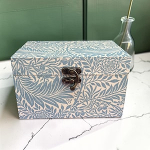 May include: A rectangular storage box featuring a light blue and white botanical leaf and flower pattern. The box has a hinged lid secured by a central antique brass-coloured metal latch. The exterior surface is covered in a repeating floral print.