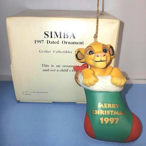 May include: A Simba Christmas ornament from 1997. The ornament features Simba, the lion cub, emerging from a green and red stocking. The stocking reads "MERRY CHRISTMAS 1997". The ornament is from Grolier Collectibles.