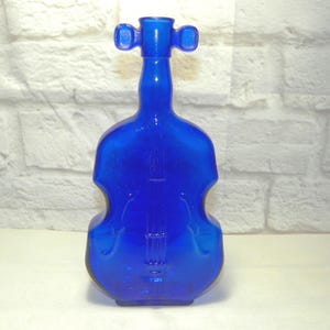 Puede incluir: Una botella de vidrio azul cobalto con forma de violín. La botella tiene un diseño detallado de violín, incluyendo las cuerdas y las efes. La parte superior tiene un diseño similar a un asa. La botella es de vidrio translúcido.