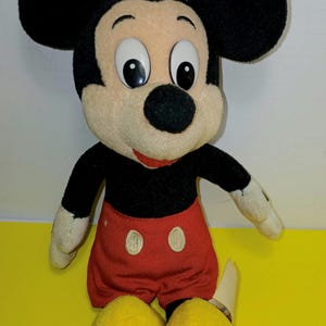 May include: A plush Mickey Mouse doll featuring large black ears, a tan face with white and black eyes, a black nose, and a red mouth. The doll wears a black shirt, red shorts with two white oval buttons, and large yellow shoes on its feet.