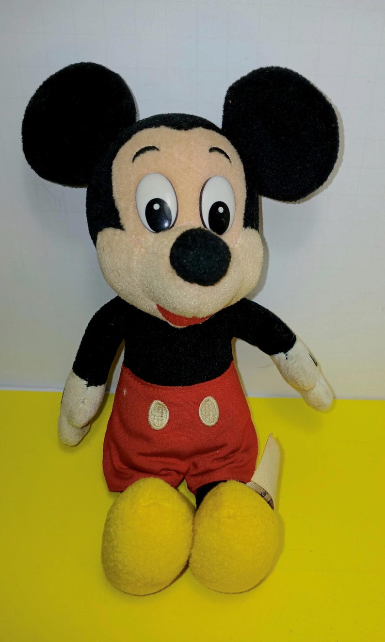 May include: A plush Mickey Mouse doll featuring large black ears, a tan face with white and black eyes, a black nose, and a red mouth. The doll wears a black shirt, red shorts with two white oval buttons, and large yellow shoes on its feet.