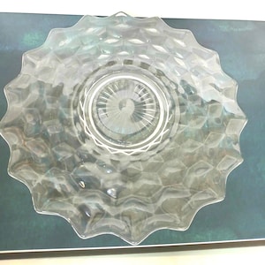 May include: Clear glass serving dish with a starburst center and a textured, geometric pattern. The rim has a scalloped edge. The dish is displayed on a dark green background.