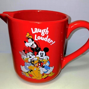May include: A bright red ceramic pitcher featuring a graphic of Disney characters including Mickey Mouse, Minnie Mouse, Donald Duck, Daisy Duck, Goofy, and Pluto. The text "Laugh Louder!" is printed in white above the characters on the front side.