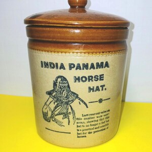 May include: A cylindrical stoneware crock with a brown lid and rim, featuring a tan body. The front displays black text reading INDIA PANAMA HORSE HAT, an illustration of a horse wearing a hat, and additional text about sales and protection.