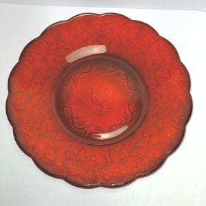 May include: A round, translucent red glass plate featuring a scalloped edge and a raised floral pattern around the rim. The center of the plate displays an embossed design of a single flower surrounded by a delicate, swirling vine motif.