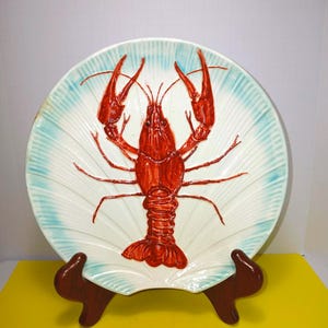 May include: A decorative ceramic plate featuring a vibrant red crawfish design. The plate has a white center with a blue rim and a fan-like pattern. The crawfish is detailed with textured elements. The plate is displayed on a brown stand.