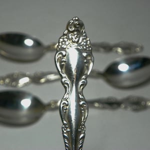 Könnte beinhalten: Nahaufnahme von vier silbernen Löffeln. Die Löffel haben verzierte Griffe mit floralen und scrollartigen Designs. Die Löffel sind fächerförmig angeordnet, wobei ein Löffel im Fokus steht. Die Löffel sind glänzend.