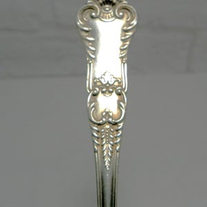 May include: Close-up of a silver spoon with ornate detailing. The spoon's handle features a decorative design with a crown-like element at the top and floral patterns. The spoon is made of a shiny, reflective metal.