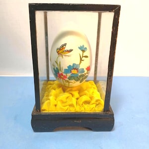Könnte beinhalten: Ein weißes, eiförmiges Ornament, verziert mit einem bunten Schmetterling sowie blauen und roten Blumenmustern, präsentiert in einer rechteckigen Glasvitrine mit schwarzem Rahmen, auf einem gelben, gerafften Stoffboden stehend.