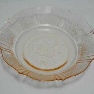 May include: A round, translucent peach-colored glass bowl featuring a scalloped edge and a decorative embossed floral pattern on the rim and center. The glass transitions from a clear center to a deeper orange hue along the outer scalloped border.