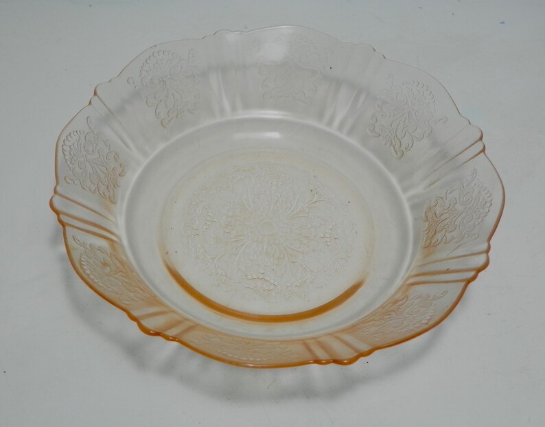 May include: A round, translucent peach-colored glass bowl featuring a scalloped edge and a decorative embossed floral pattern on the rim and center. The glass transitions from a clear center to a deeper orange hue along the outer scalloped border.
