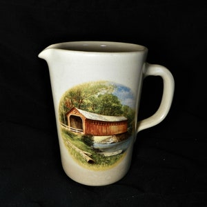 May include: Cream ceramic pitcher with a handle and spout. The pitcher has a painted oval scene of a covered bridge over a river, surrounded by trees. The bridge is painted in shades of red and brown.
