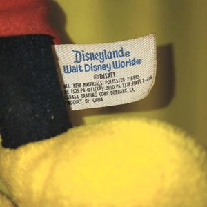 May include: A plush toy with a red fabric section and a yellow fabric section, featuring a white fabric tag printed with Disneyland, Walt Disney World, Disney, and manufacturing details including polyester fibers and product of China.