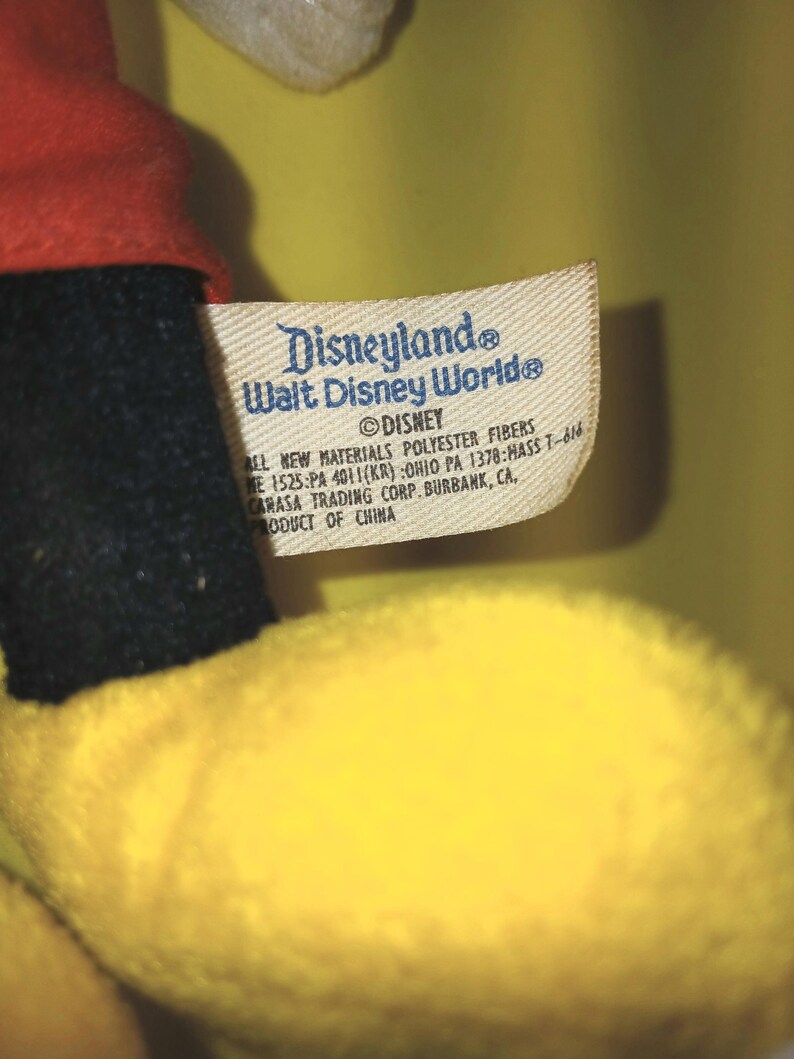 May include: A plush toy with a red fabric section and a yellow fabric section, featuring a white fabric tag printed with Disneyland, Walt Disney World, Disney, and manufacturing details including polyester fibers and product of China.