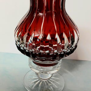 Pode incluir: Um castiçal de vidro vermelho rubi com uma base de vidro transparente. A parte superior tem um design facetado e a base é redonda com um padrão de estrela. O castiçal tem aproximadamente 15 cm de altura.