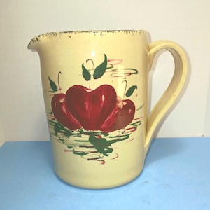 May include: A cream-colored ceramic pitcher with a large handle and a spout. The pitcher is decorated with three red apples and green leaves. The rim has a distressed, dark green trim.