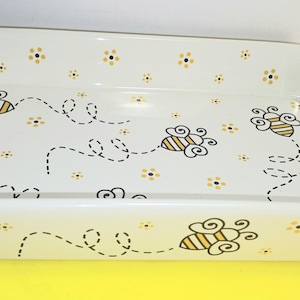 Könnte beinhalten: Rechteckige Keramik-Backform mit weißem Hintergrund, verziert mit gelben und schwarzen Bienen und kleinen gelben Blumen. Die Bienen fliegen entlang gestrichelter Linien. Die Form hat abgerundete Ecken und einen leicht erhöhten Rand.