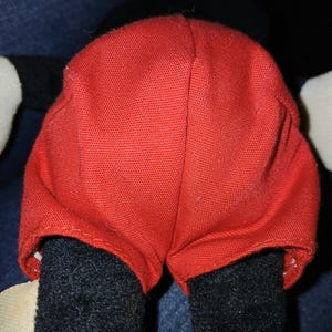 May include: A plush toy featuring bright red fabric shorts with a visible woven texture. The shorts are worn over black fabric legs, with white fabric visible at the sides. The construction shows a central vertical seam and stitched hems.