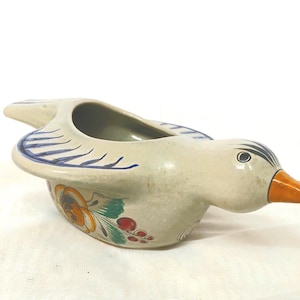 Könnte beinhalten: Ein keramischer, vogelförmiger Pflanztopf mit cremefarbenem Körper, blauen Flügelakzenten und einem orangefarbenen Schnabel. Der Pflanztopf hat ein Blumendesign mit orangefarbenen, gelben und grünen Details. Der Vogel hat eine ausgehöhlte Pflanzfläche.